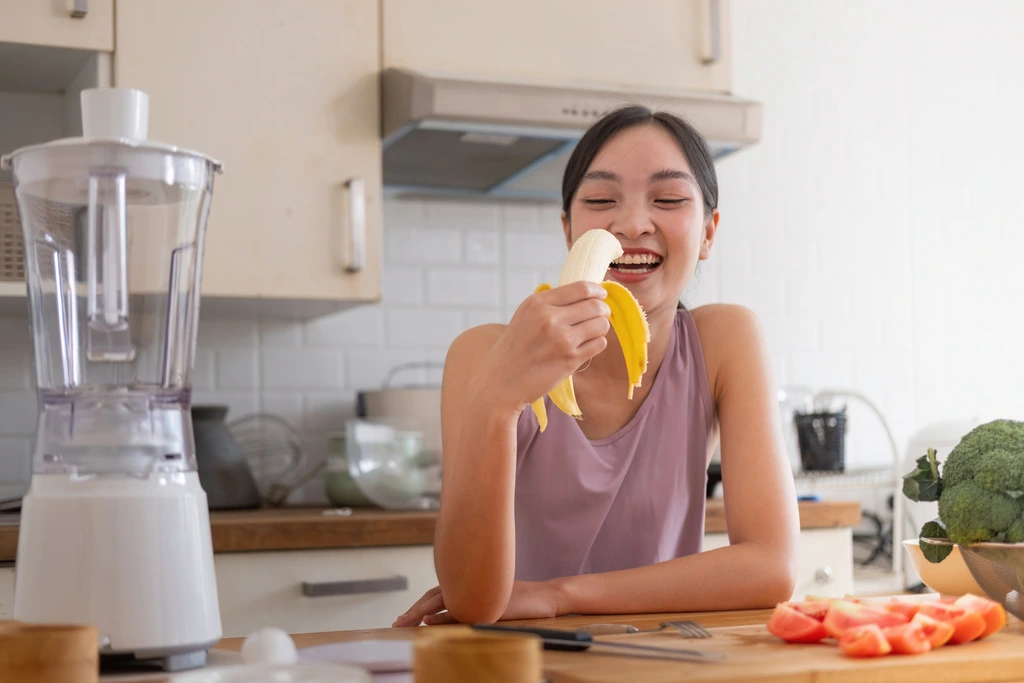 Juliana Cruz of Alchemy Chef laughing while eating a fresh banana at her kitchen counter, with a blender and other ingredients nearby.