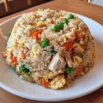 A dome-shaped serving of flavorful chicken fried rice mixed with visible chunks of chicken, peas, carrots, and scrambled egg.
