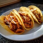 Three corn tortilla chicken tacos filled with shredded, saucy meat and topped with white cheese.