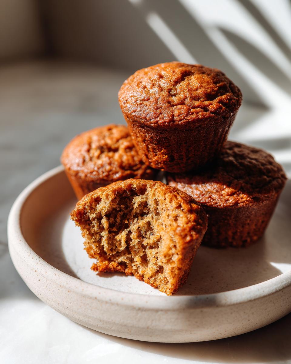 A stack of four golden-brown banana protein muffins, one broken open to show the moist interior.