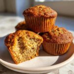 A stack of golden brown banana protein muffins, with one muffin broken open to show the moist interior texture.