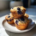 A stack of three golden-brown blueberry protein muffins on a white plate, highlighted by bright sunlight.