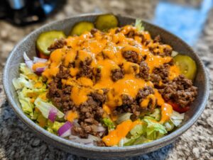 A close-up view of a hearty cheeseburger bowl featuring seasoned ground beef, orange sauce, lettuce, and pickle slices.