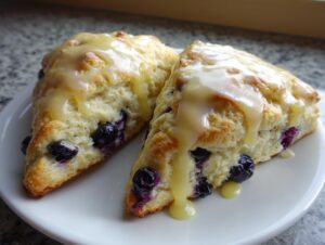 Two triangular blueberry scones generously topped with a thick, dripping lemon glaze, served on a white plate.