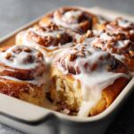 Close-up of fluffy cinnamon roll fresh from the oven, covered in thick white icing and chopped pecans.