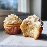 Two fluffy vanilla cupcakes, one whole and one bitten open to show the moist crumb texture, topped with white frosting.