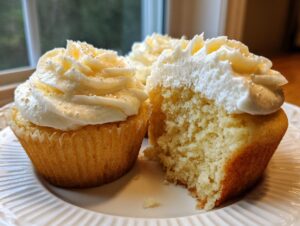Close-up of fluffy vanilla cupcakes, one bitten to show the moist crumb, topped with white frosting and sprinkles.