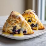 Two triangular blueberry scones generously topped with a thick, sweet yellow glaze, sitting on a white plate.