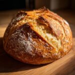 A close-up of a round, golden-brown loaf of homemade french bread with a beautifully scored, cracked crust dusted with flour.