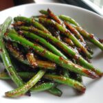 A close-up of vibrant green beans coated in a glossy, dark honey garlic sauce.