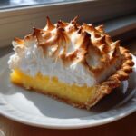 A close-up slice of bright yellow lemon meringue pie with tall, perfectly browned meringue peaks.