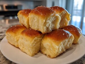 A stack of six fluffy, golden-brown dinner rolls brushed with a shiny glaze, resting on a white plate.