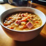 A close-up of a steaming white bowl filled with thick, hearty ham and bean soup featuring chunks of ham and carrots.