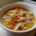 Close-up of a white bowl filled with rich turkey soup, featuring shredded turkey, egg noodles, carrots, and celery.