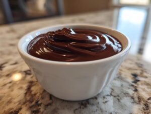 Close-up of a small white bowl filled with glossy, rich chocolate dip, sitting on a granite countertop.