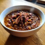 A close-up of a steaming white bowl filled with rich, hearty 15-bean soup topped with shredded pulled pork.