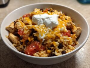 A close-up view of an amazing chicken burrito bowl featuring seasoned rice, black beans, diced tomatoes, melted cheese, and a dollop of sour cream.