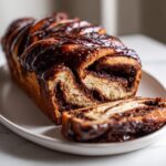 A freshly baked chocolate babka loaf, drizzled with rich chocolate glaze, with one slice cut and resting beside it.