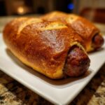 Close-up of two freshly baked pretzel dogs with shiny, dark brown crusts on a white rectangular plate.