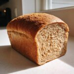 A golden brown loaf of homemade whole wheat bread sitting on a counter, with one end sliced to show the soft interior texture.