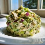 A perfectly molded scoop of creamy avocado chicken salad featuring chunks of chicken, red onion, and celery, served on a white plate.