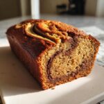 A golden-brown, freshly baked loaf of banana zucchini bread with a visible swirl pattern and banana slice on top.