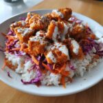 A close-up of a white bowl filled with Bang Bang Salmon Bowls, featuring crispy salmon pieces over rice and slaw, drizzled with white sauce.