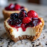 A close-up of a berry-topped mini cheesecake with a bite taken out, showing the creamy filling and graham cracker crust.