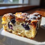 A close-up of a baked slice of blueberry french toast casserole featuring a creamy center and powdered sugar dusting.