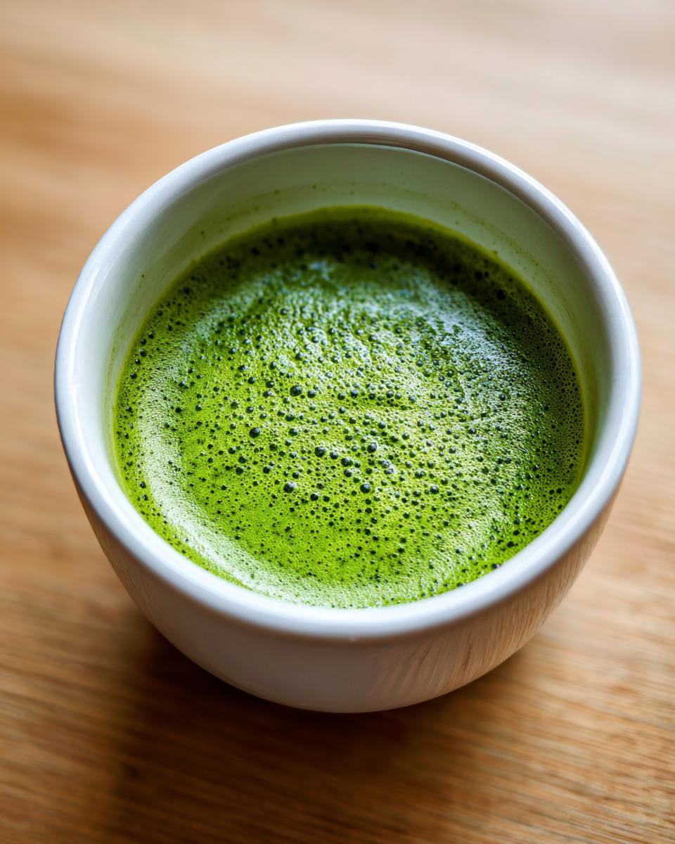 Close-up of a vibrant green, frothy beverage, possibly matcha, in a white bowl, contrasting with a wooden surface.