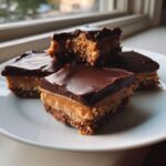 Four squares of rich buckeye fudge with a peanut butter layer and chocolate topping stacked on a white plate.