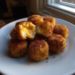 A pile of golden brown, crispy potato croquettes on a white plate, one is broken open showing melted cheese inside.