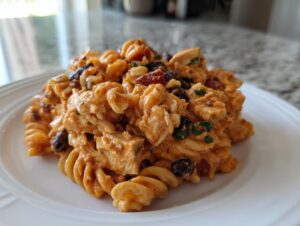 A close-up serving of creamy, cheesy enchilada pasta mixed with chunks of chicken and black beans on a white plate.