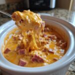 A spoonful of hot, cheesy Reuben dip showing a dramatic cheese pull over the crockpot.