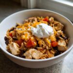 Close-up of a white bowl filled with a chicken burrito bowl featuring seasoned rice, chicken chunks, black beans, tomatoes, melted cheese, and sour cream.