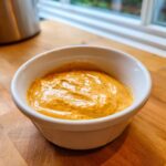 Close-up of creamy, orange-colored beer cheese dip served in a small white ramekin on a wooden surface.