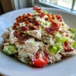 Close-up of a creamy blt chicken salad topped generously with crispy bacon bits and a cherry tomato wedge.