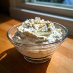 A clear glass bowl filled with creamy, thick blue cheese dip, topped with extra crumbles.