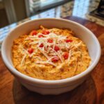 A close-up of a creamy orange boat dip in a white bowl, topped with shredded white cheese and diced red peppers.