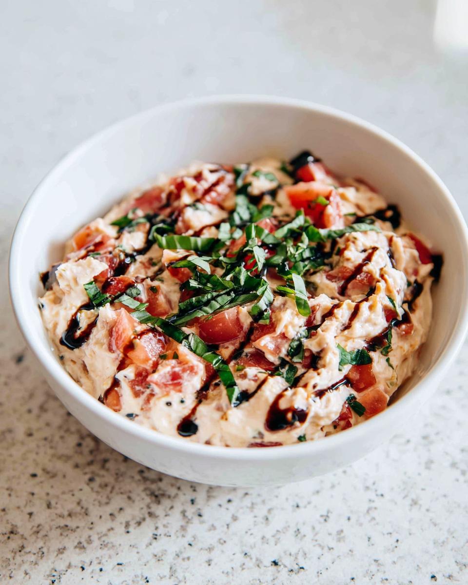 A close-up of creamy bruschetta dip topped with diced tomatoes, fresh basil, and a balsamic glaze drizzle.