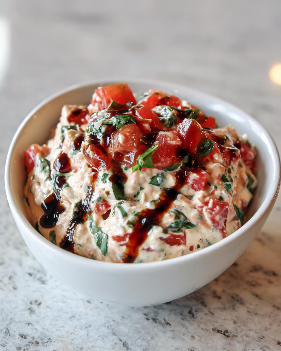 A close-up of creamy bruschetta dip mixed with tomatoes and basil, drizzled with balsamic glaze in a white bowl.
