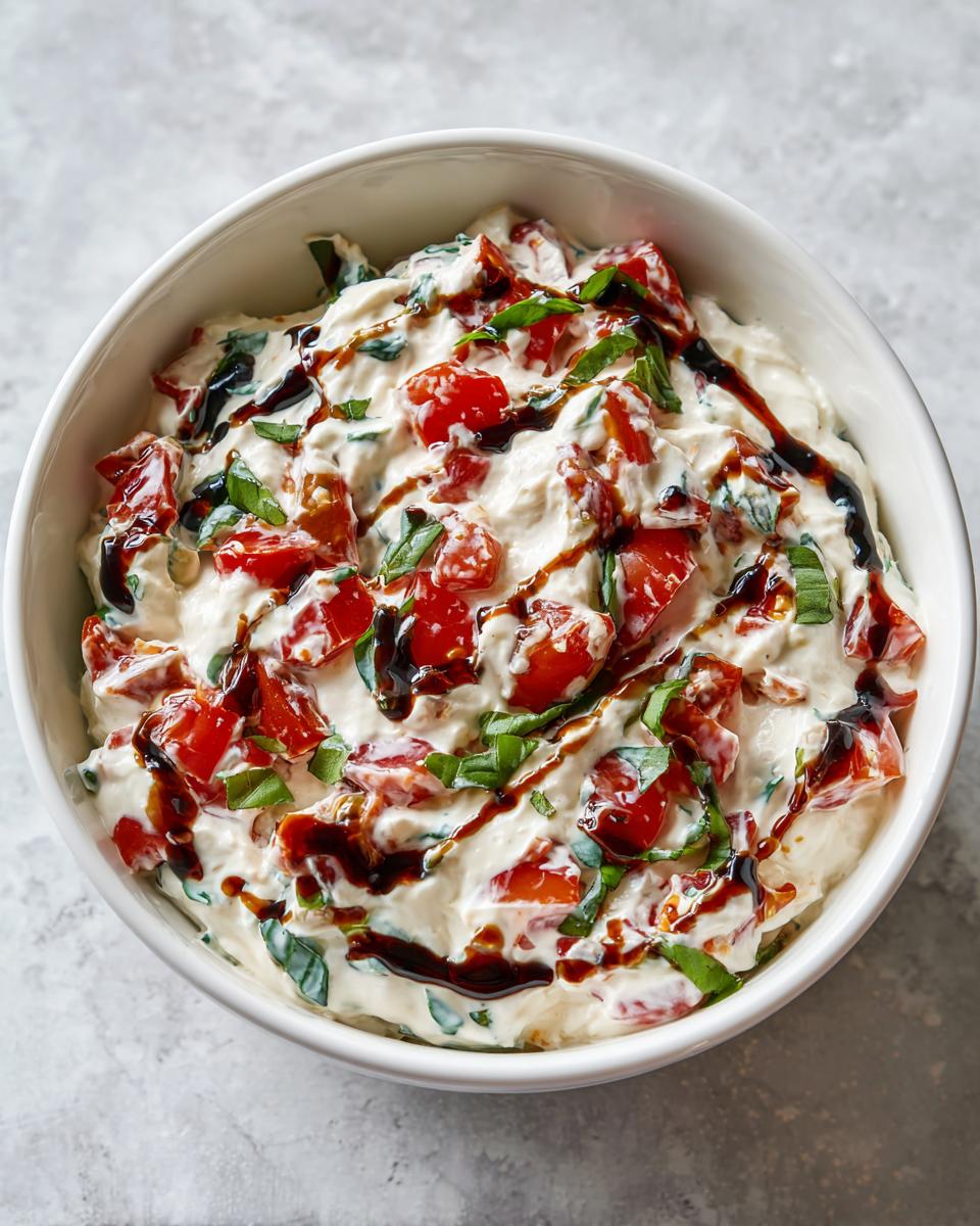 Close-up of creamy bruschetta dip topped with diced tomatoes, fresh basil, and a balsamic glaze drizzle.