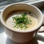 A close-up of creamy cauliflower soup in a white mug, topped generously with fresh chopped chives and pepper.