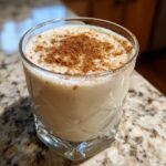 Close-up of a glass filled with creamy, light-colored coquito, dusted generously with ground cinnamon.