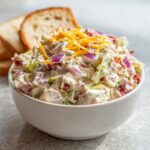 A white bowl filled with creamy hoagie dip topped with shredded cheese and red onion, served next to slices of toasted bread.