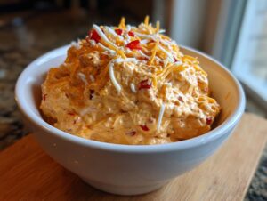 A close-up of a white bowl filled with creamy orange boat dip, topped with shredded cheddar and white cheese.