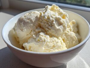 A white bowl filled with scoops of creamy, pale yellow protein ice cream, brightly lit by sunlight.