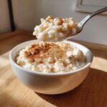 A spoonful of creamy rice pudding recipe topped with cinnamon being lifted from a white bowl.