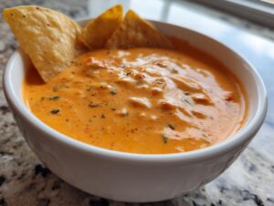 Close-up of a white bowl filled with creamy, orange rotel dip garnished with herbs, served with tortilla chips.