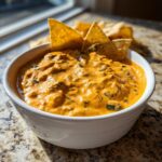 A close-up of creamy, orange-colored rotel dip in a white bowl, garnished with tortilla chips for dipping.